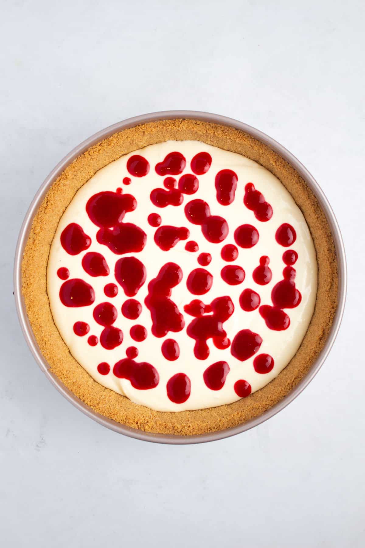 Circular baking pan full of crust and cheesecake batter with splashes of sauce on the top.