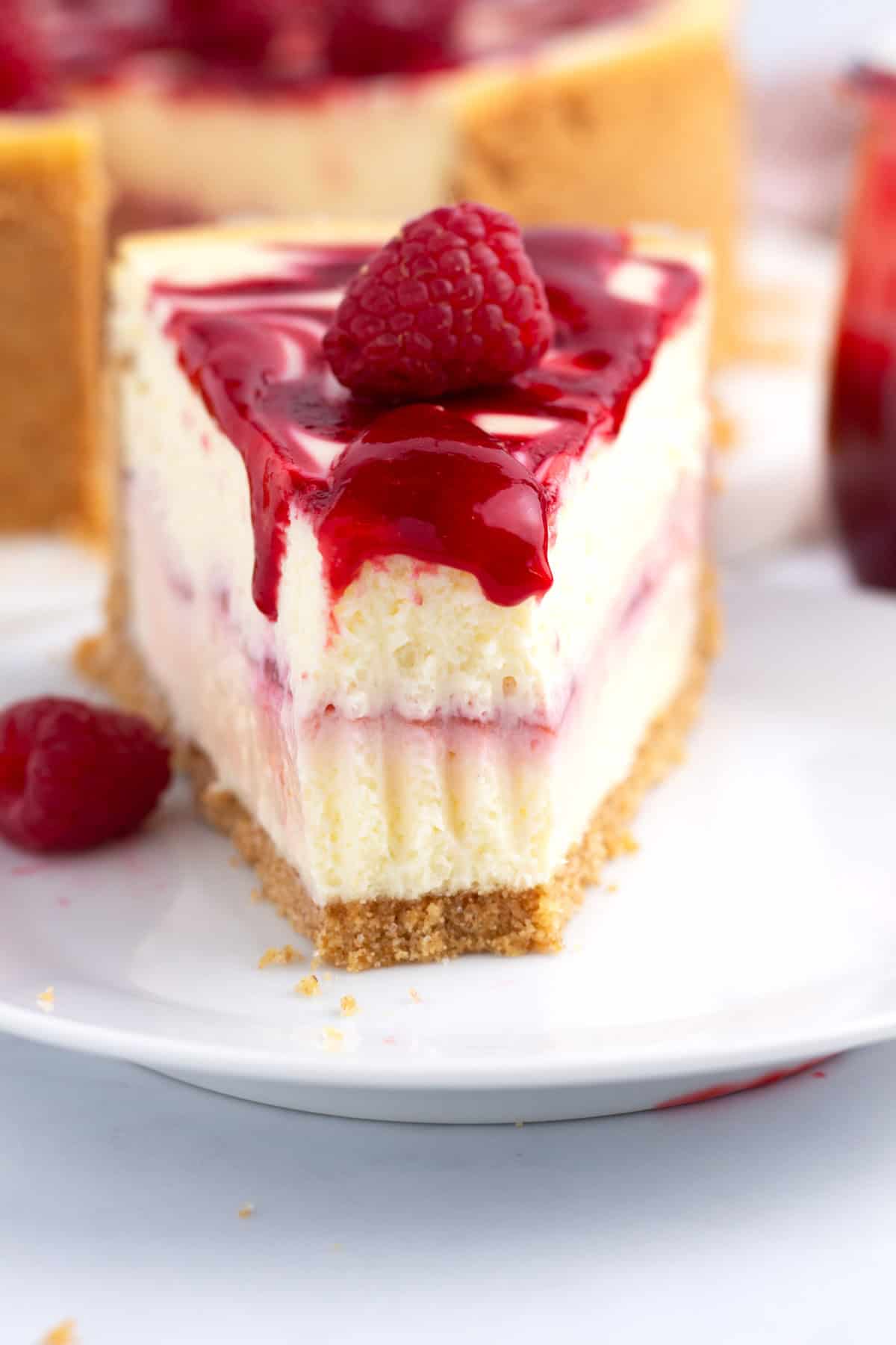 A slice of cheesecake on a plate with topping and raspberries on it and beside it.