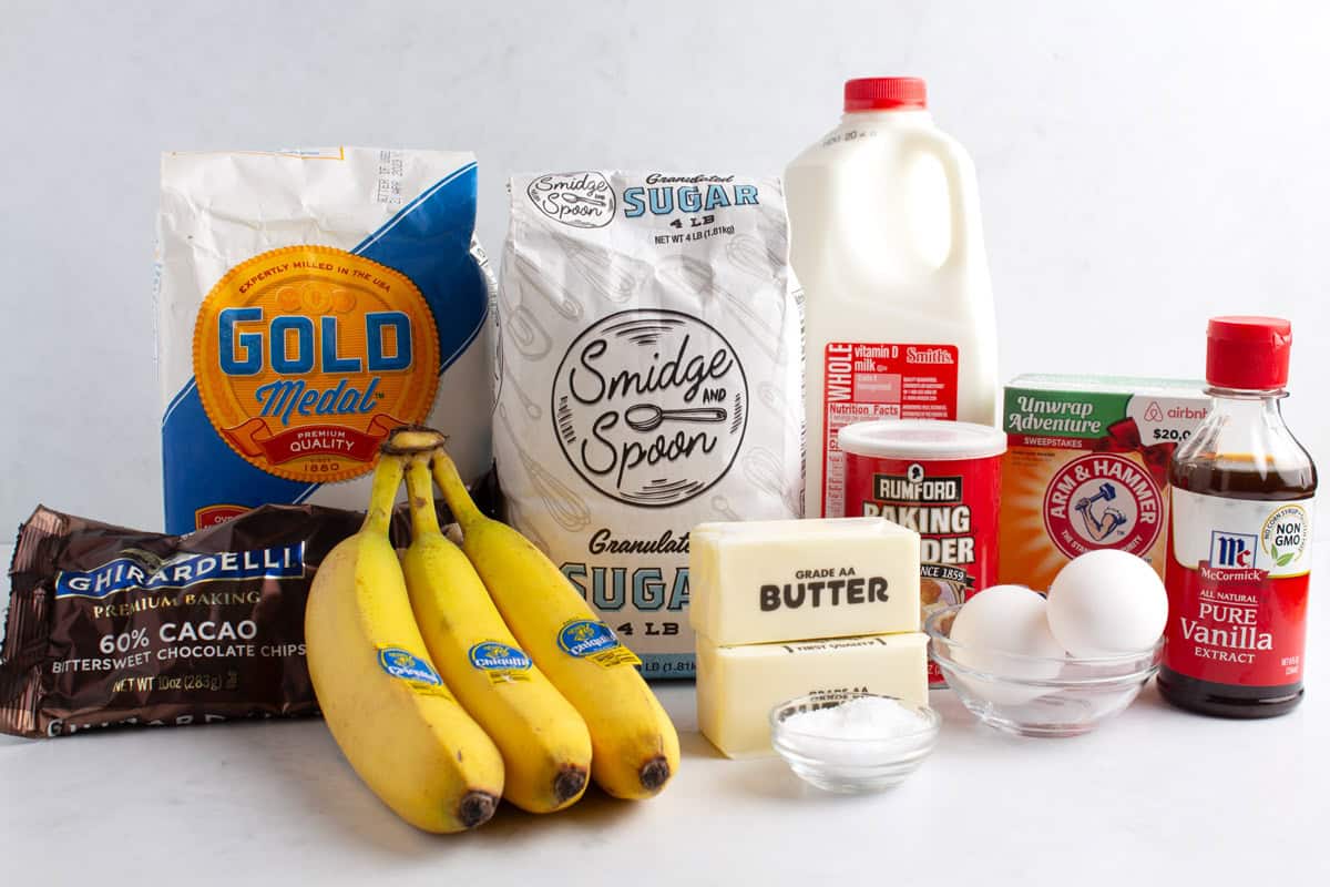 Flour, bananas, chocolate chips, sugar, butter, a small dish of salt, eggs, baking powder, baking soda, milk, and vanilla extract on a countertop.