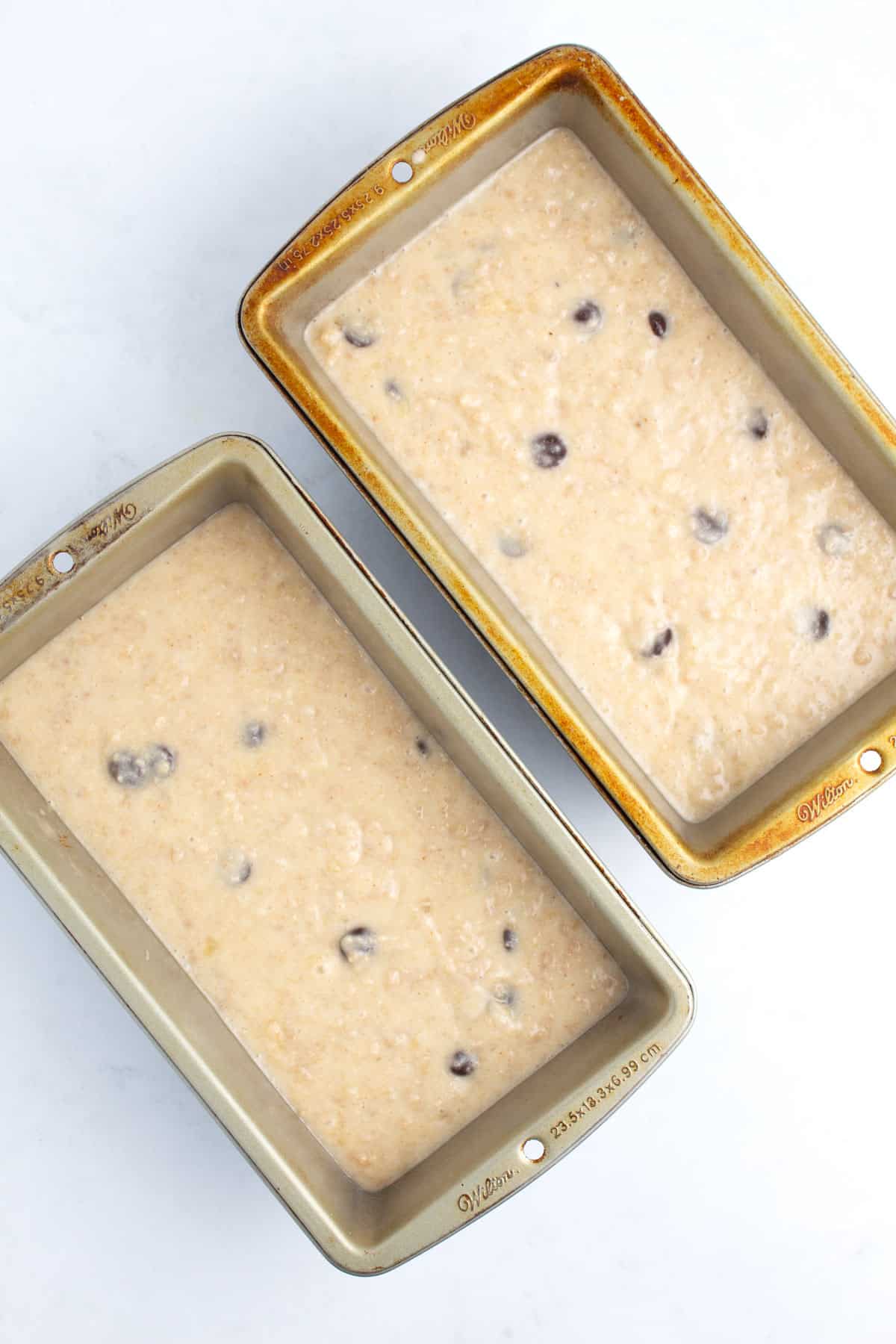 Two bread baking pans with bread dough in them on a white surface.