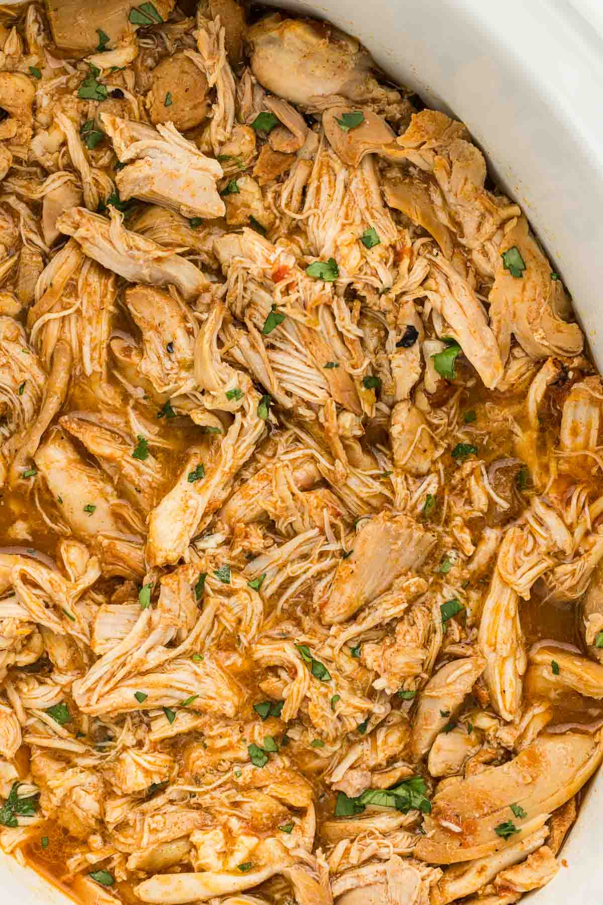 Shredded Mexican chicken floating in juices in a white crockpot.