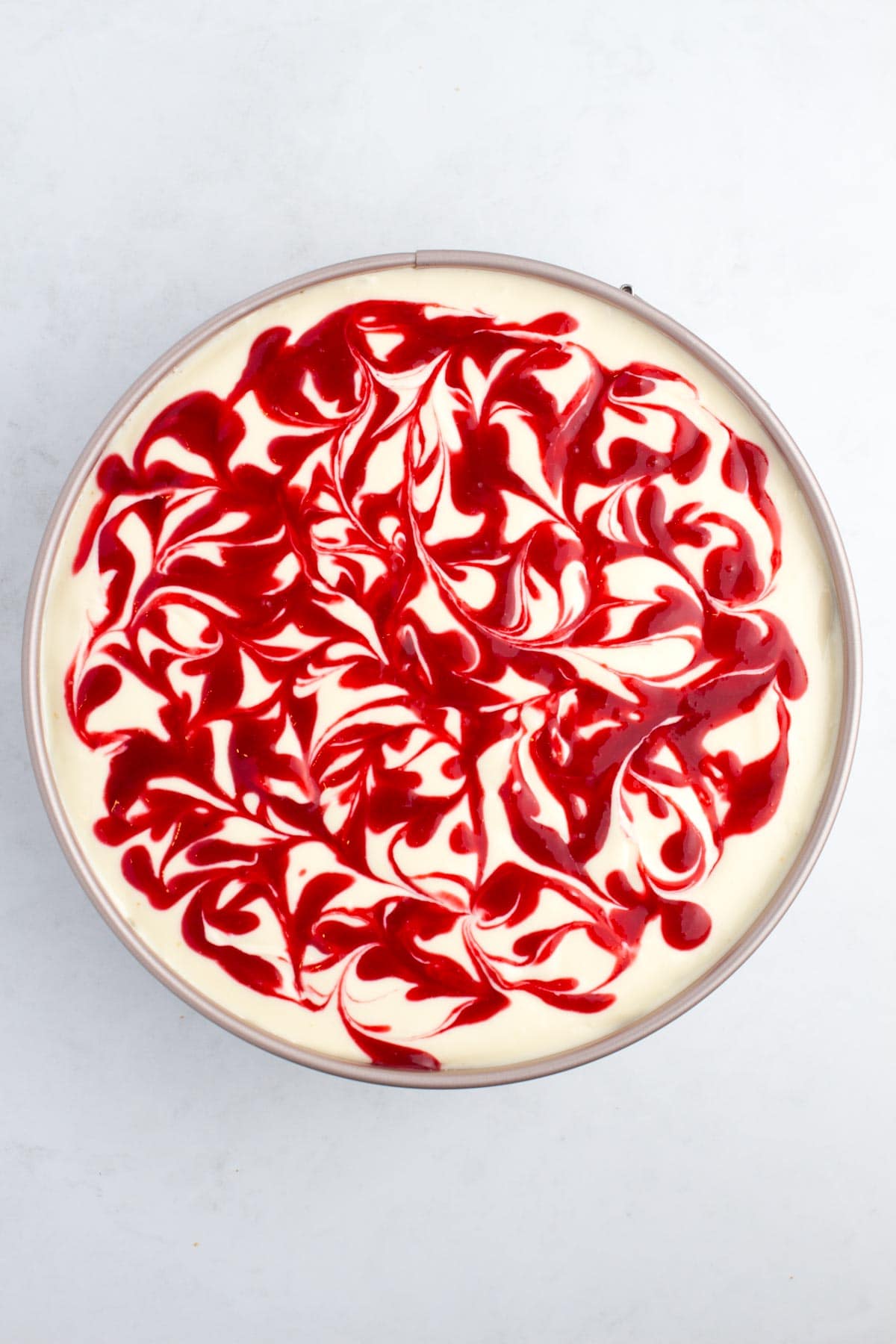 Uncooked cheesecake in a pan with raspberry sauce swirled in the top.