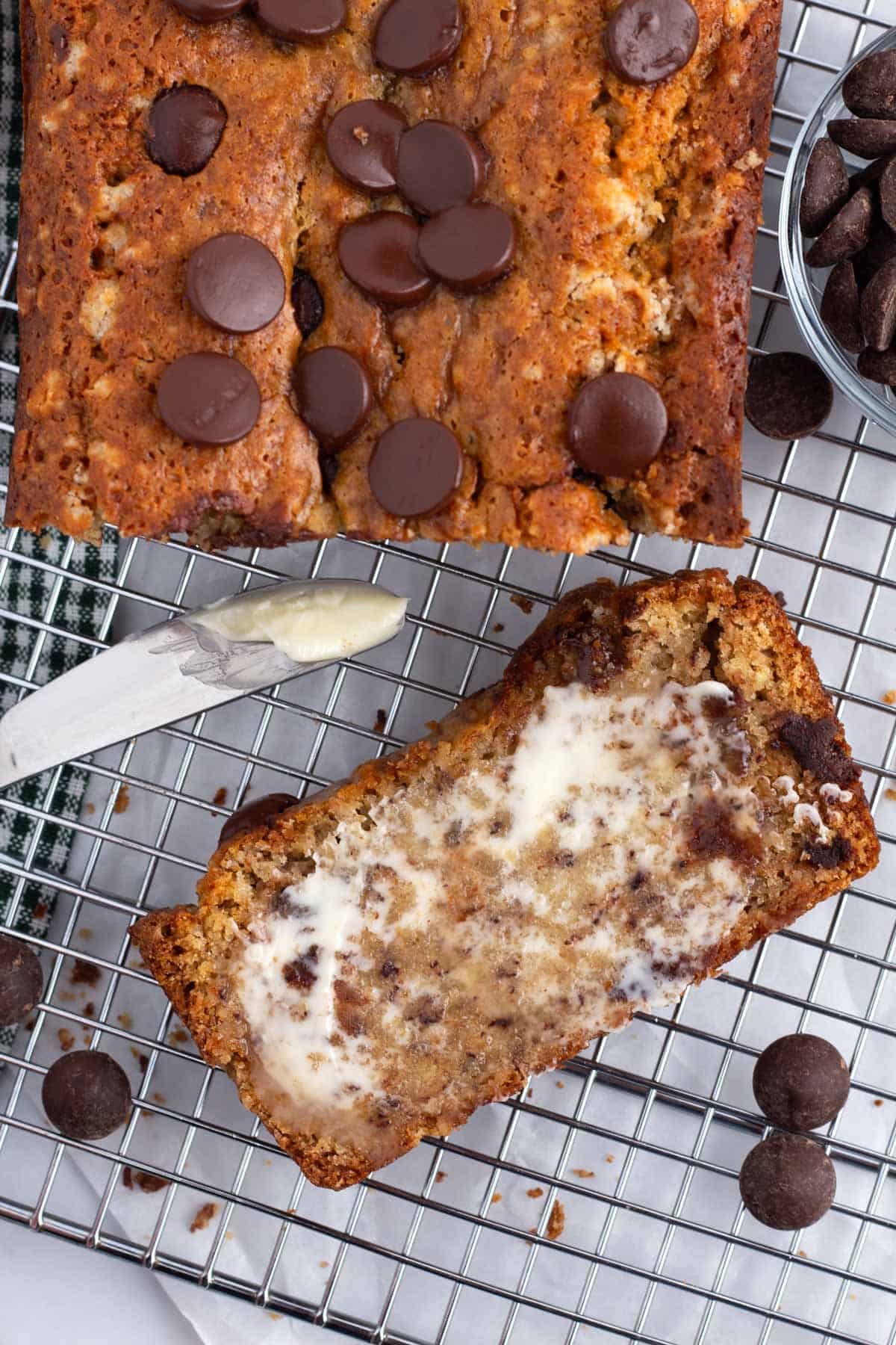 A slice of banana bread on a cooling rack next to a loaf and a butter knife.