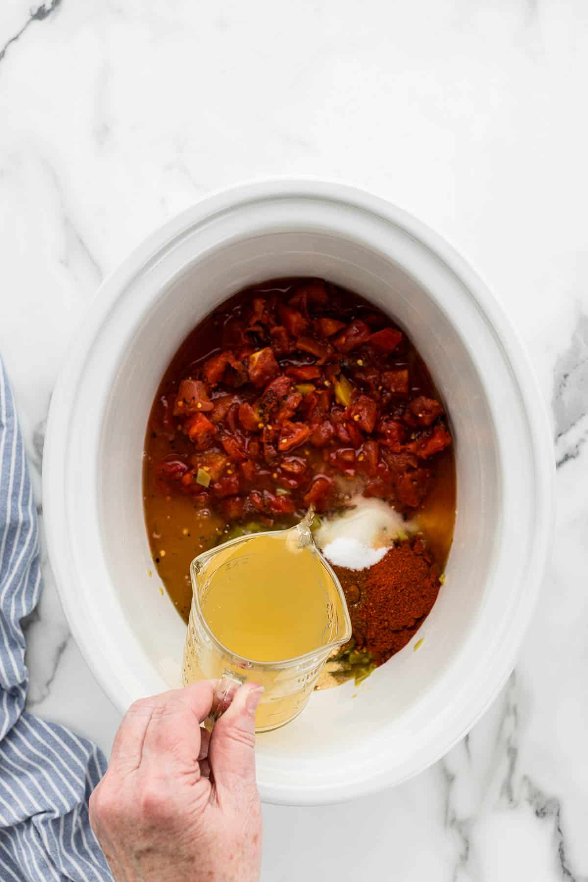 A white slow cooker full of ingredients with a measuring cup of chicken broth getting poured into it.