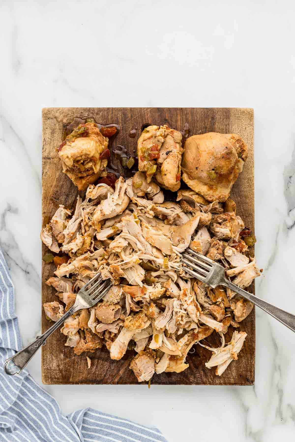 A wooden cutting board with shredded chicken and two upside-down forks on it.