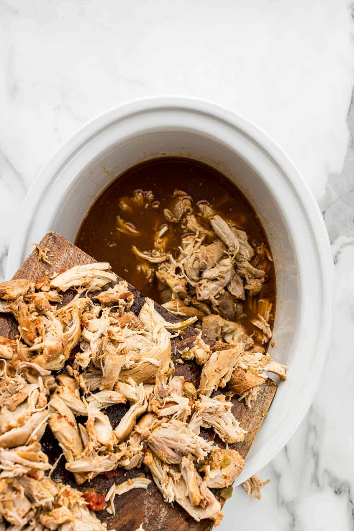 A cutting board with shredded chicken getting poured into a white crockpot.