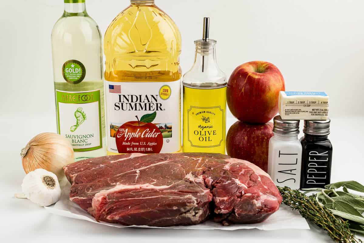Garlic, an onion, meat, apple cider, olive oil, apples, salt and pepper, butter, herbs, and white wine on a white surface.