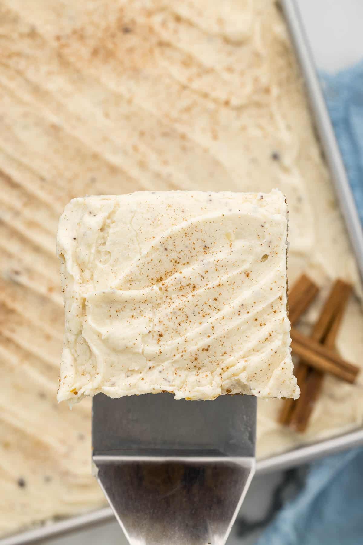 A serving tool holding a piece of cake with cream cheese frosting over a baking pan of a whole cake.