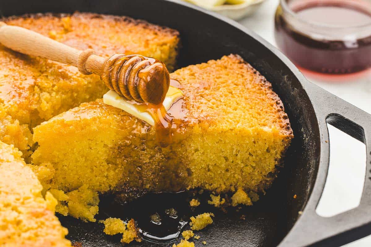 Sliced cornbread in a skillet with butter and hot honey drizzled over a piece.