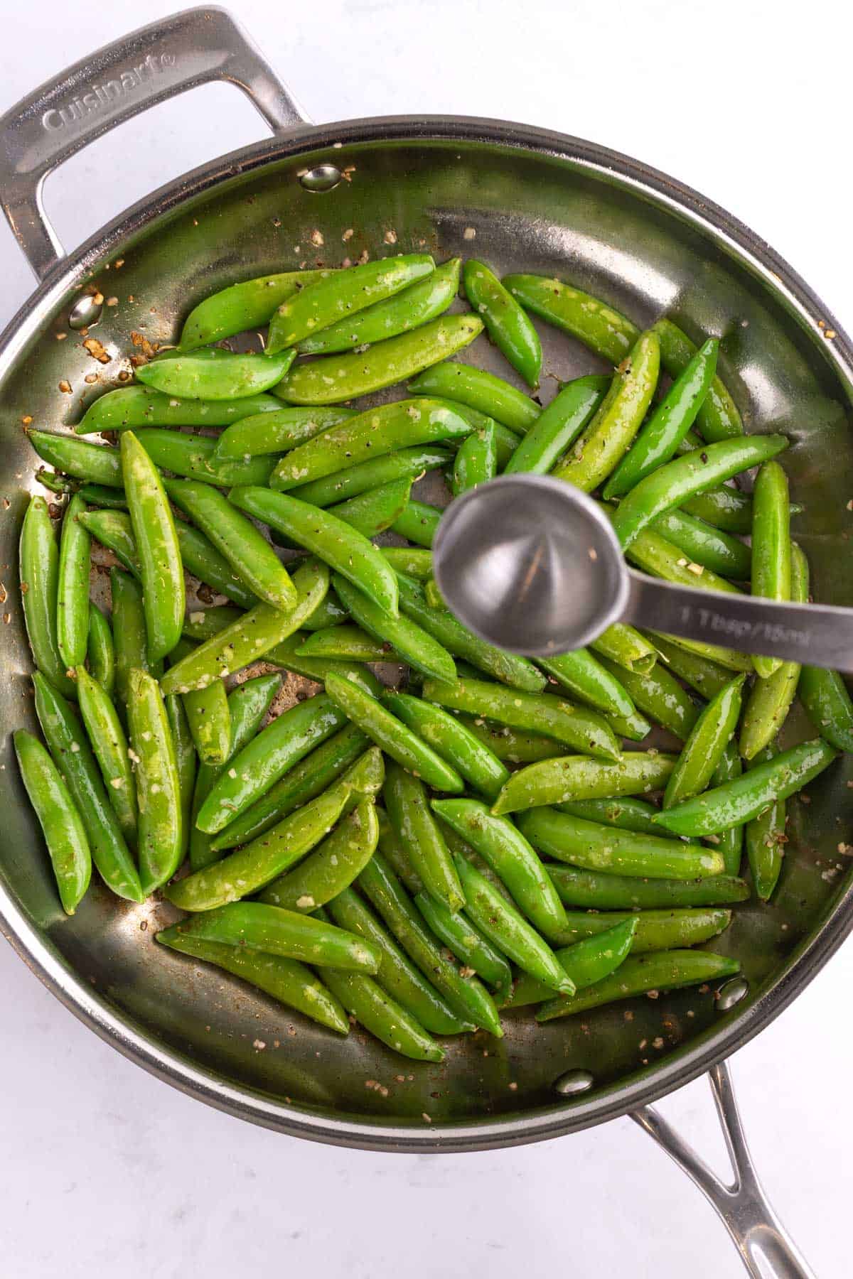 A measuring spoon over a cooking pan of pea pods.