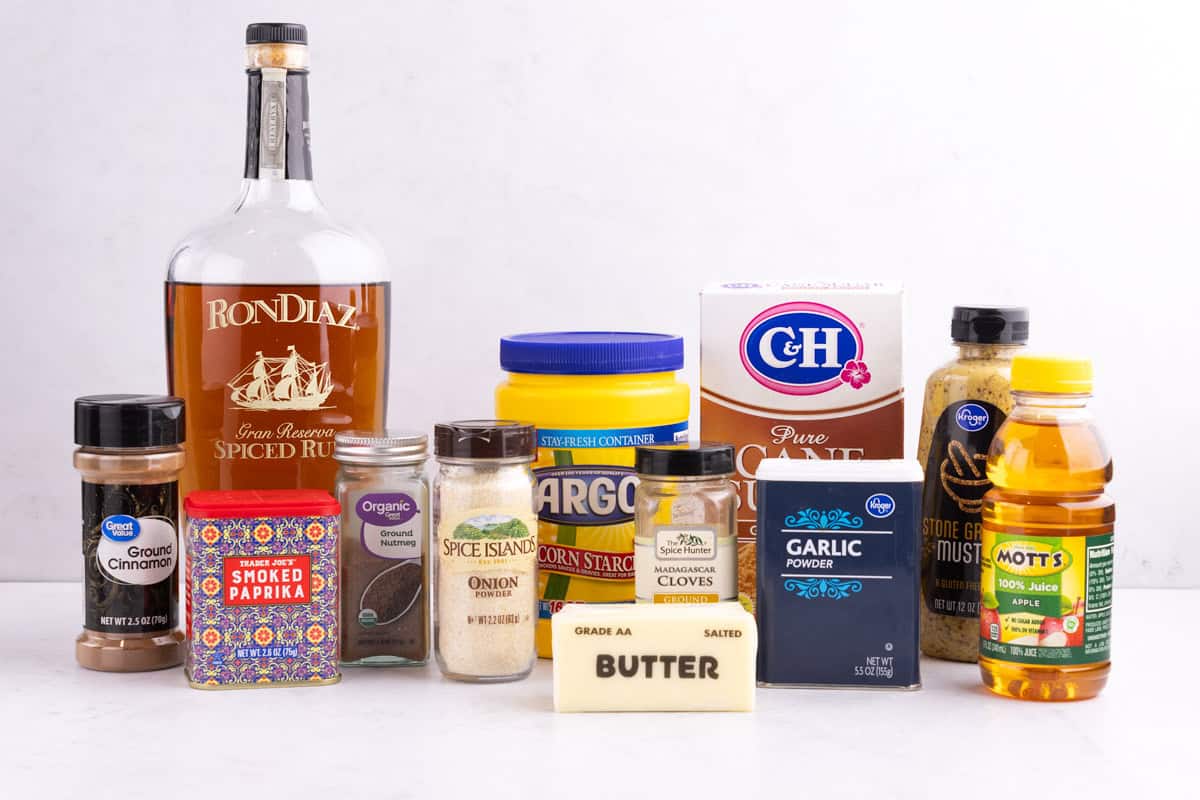 Cinnamon, paprika, spiced rum, nutmeg, onion powder, butter, corn starch, cloves, cane sugar, garlic powder, apple juice, and ground mustard on a counter.