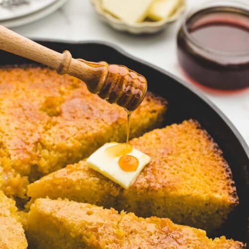 A honey stick drizzling spicy honey over a piece of cornbread in a skillet.