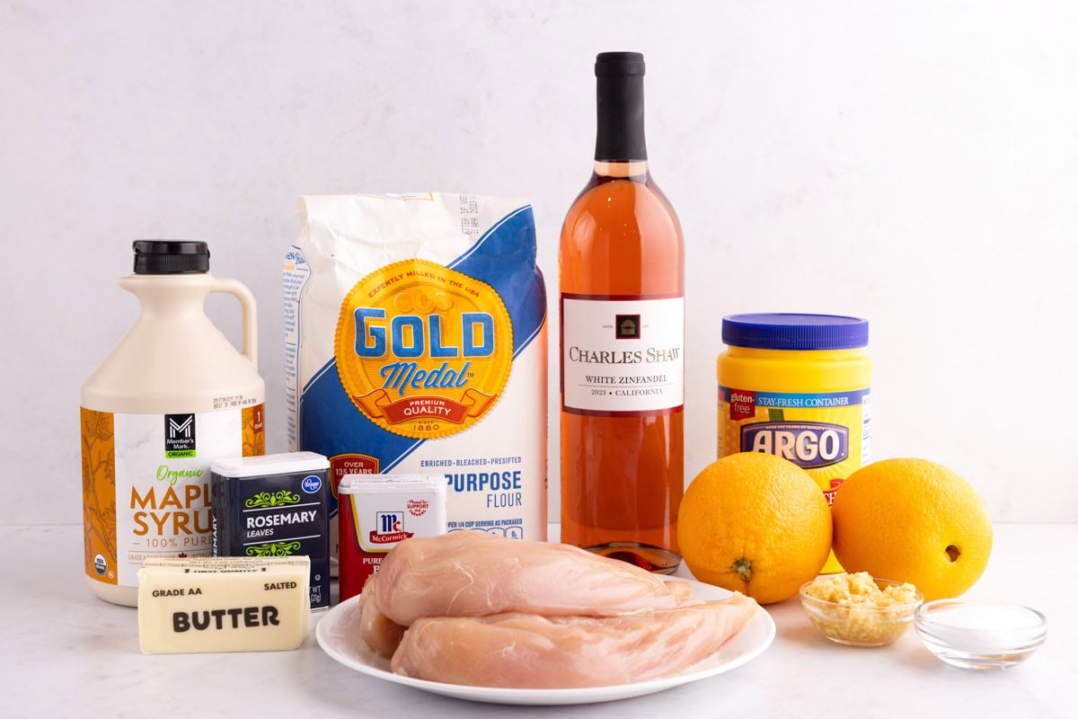 Maple syrup, butter, seasonings, flour, oranges, garlic, salt, chicken breasts, white wine, and corn starch on a white surface.