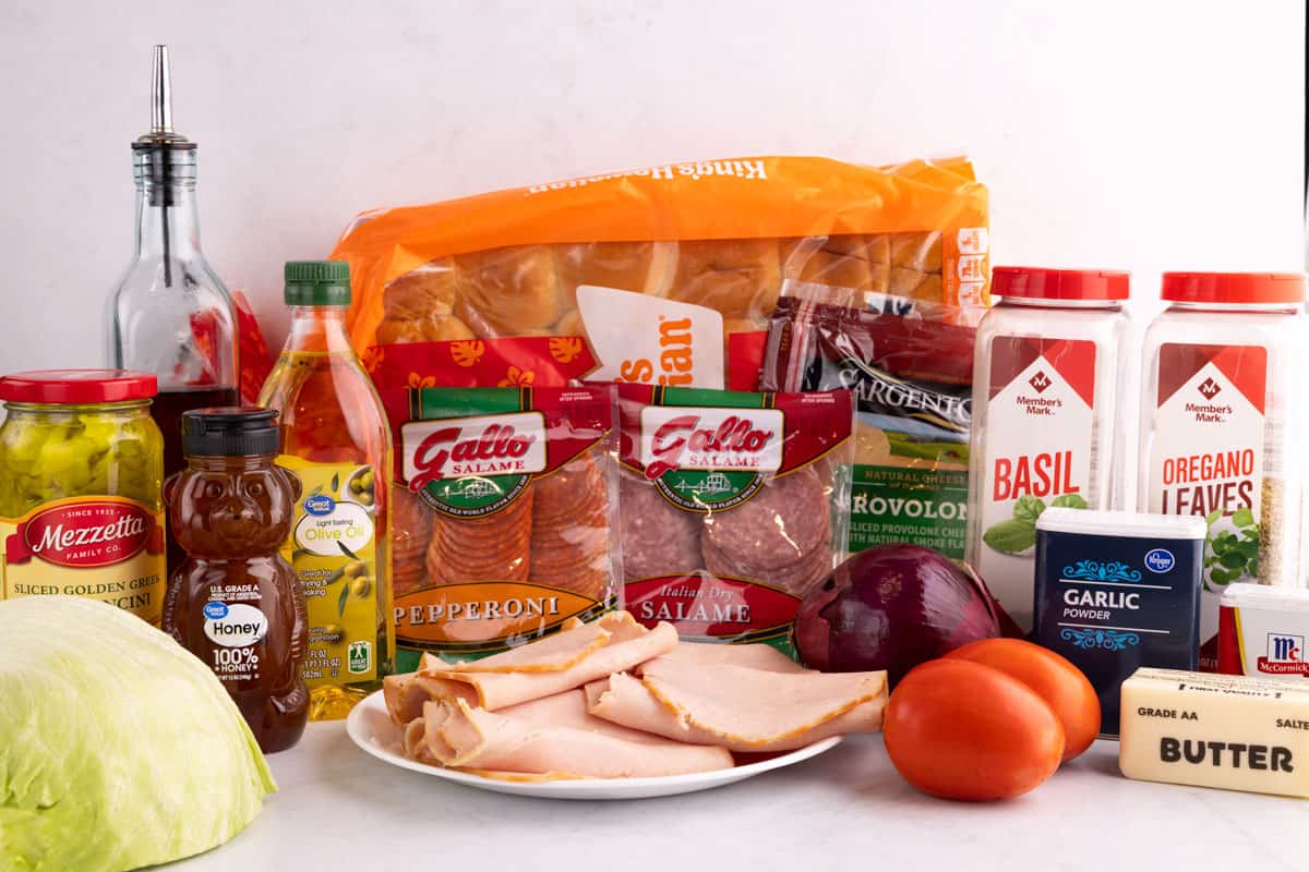 Seasonings, salami, lettuce, Hawaiian rolls, turkey, pepperoni, provolone, onion, tomatoes, butter, honey, oil, vinegar, and pepperoncini on a counter.