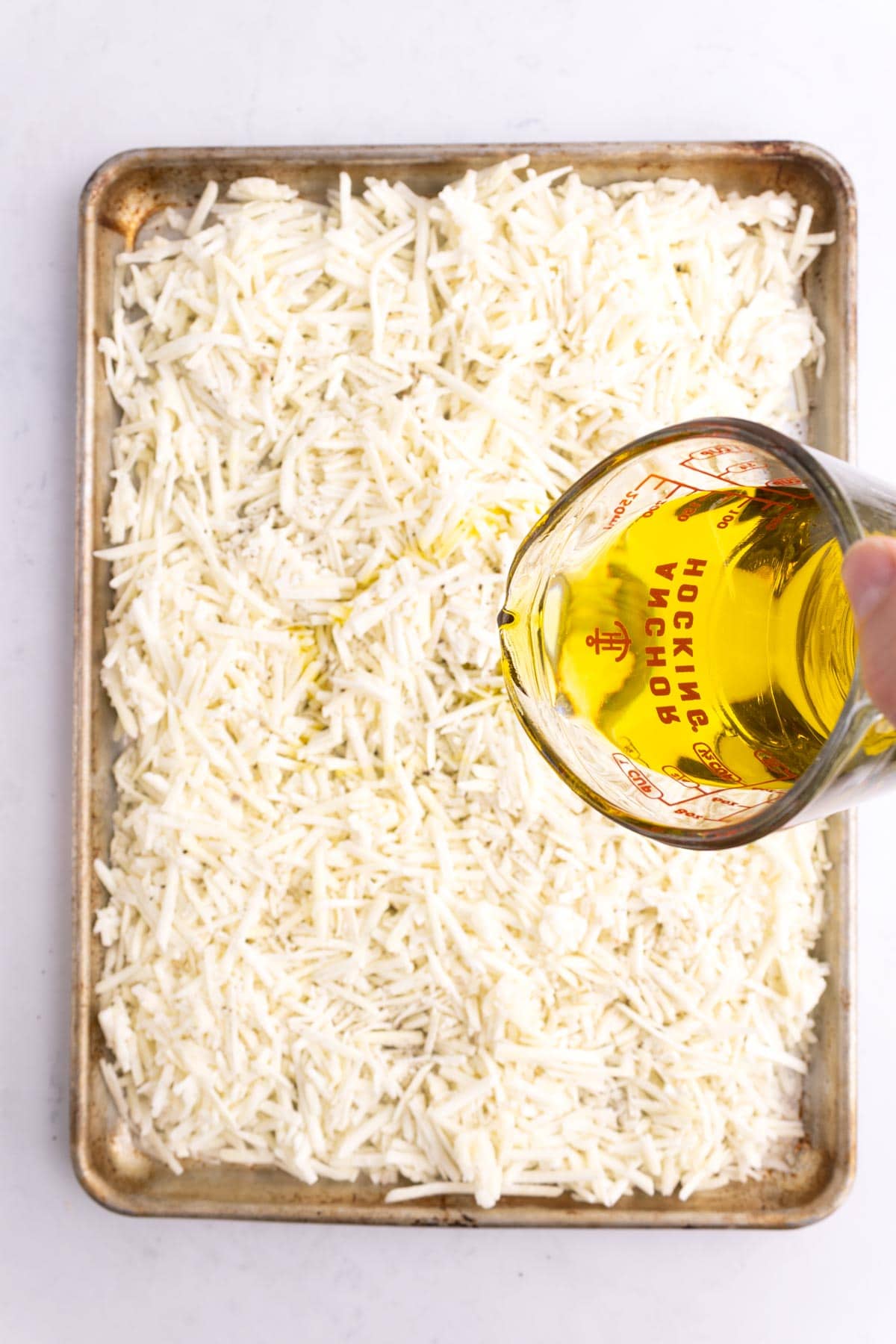 A measuring dish pouring oil onto a pan of frozen potatoes.