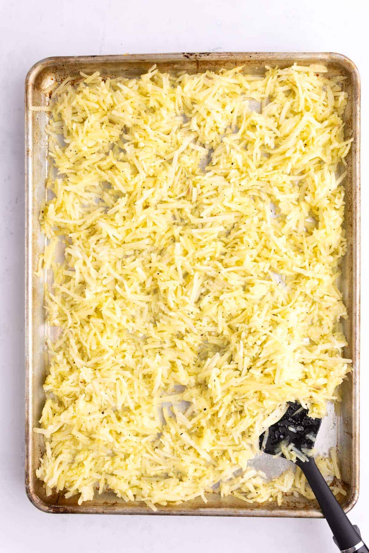 Oil mixed into frozen potatoes with a rubber spatula on a sheet pan.