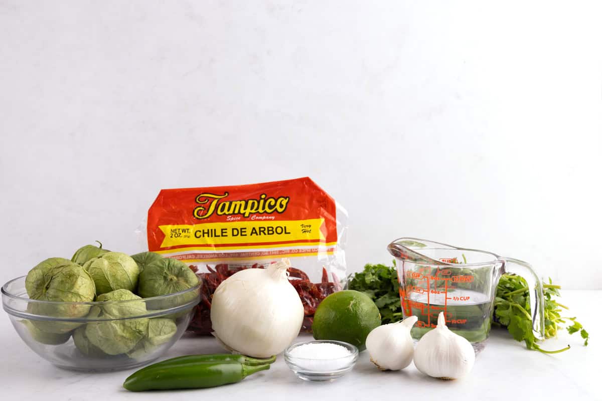 Tomatillos, garlic, onion, chile de arbol, peppers, cilantro, salt, water, and cloves on a white countertop.