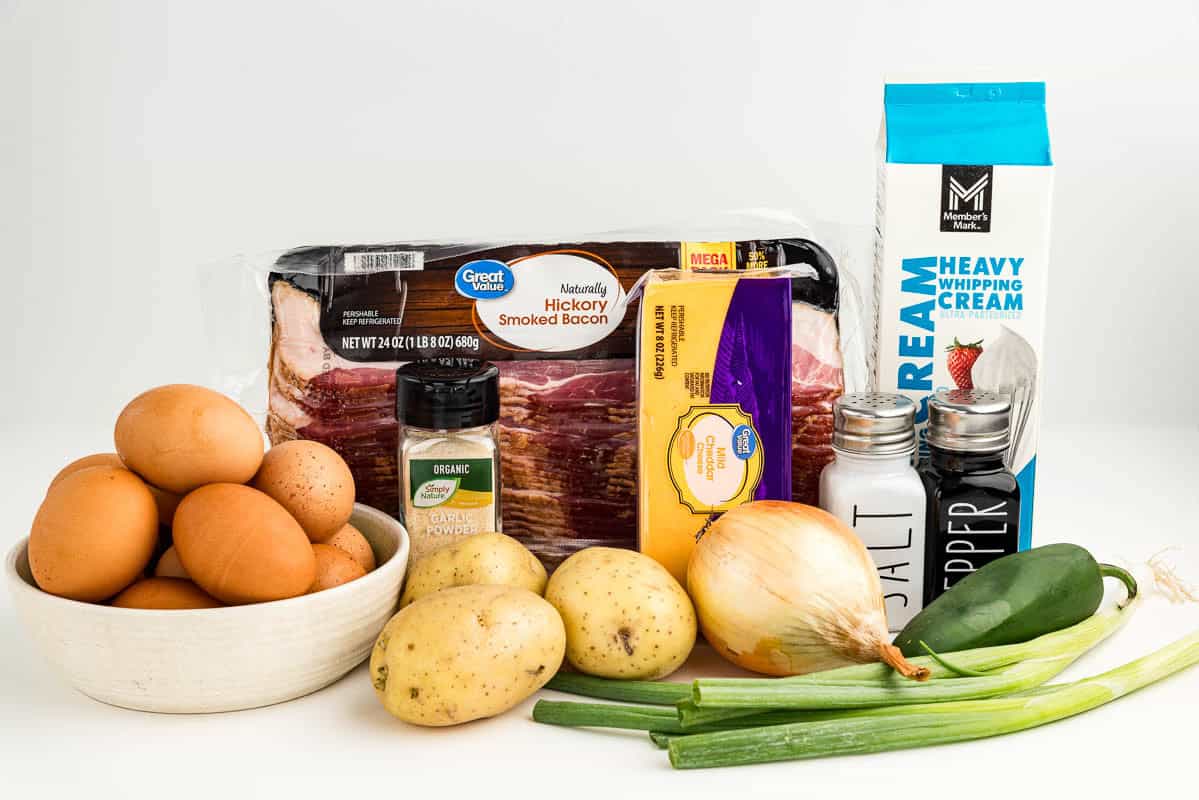 Eggs, potatoes, onion, salt and pepper, heavy cream, cheese, garlic powder, bacon, a jalapeno, and green onions on a counter.