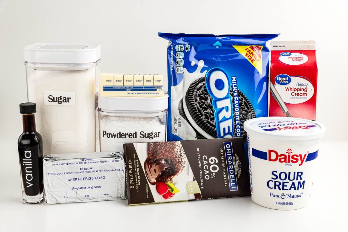 Vanilla, sugar, butter, powdered sugar, cream cheese, oreos, chocolate, heavy cream, and sour cream on a countertop.
