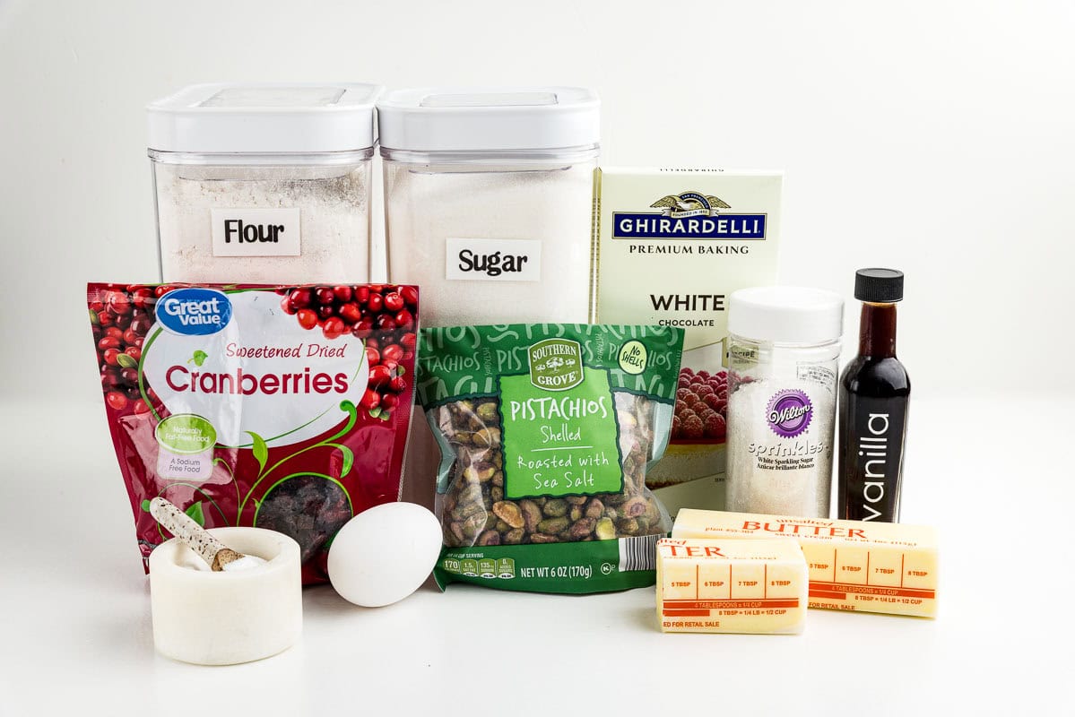 Salt, an egg, butter, vanilla, sprinkles, cranberries, pistachios, flour, sugar, and white chocolate on a counter.