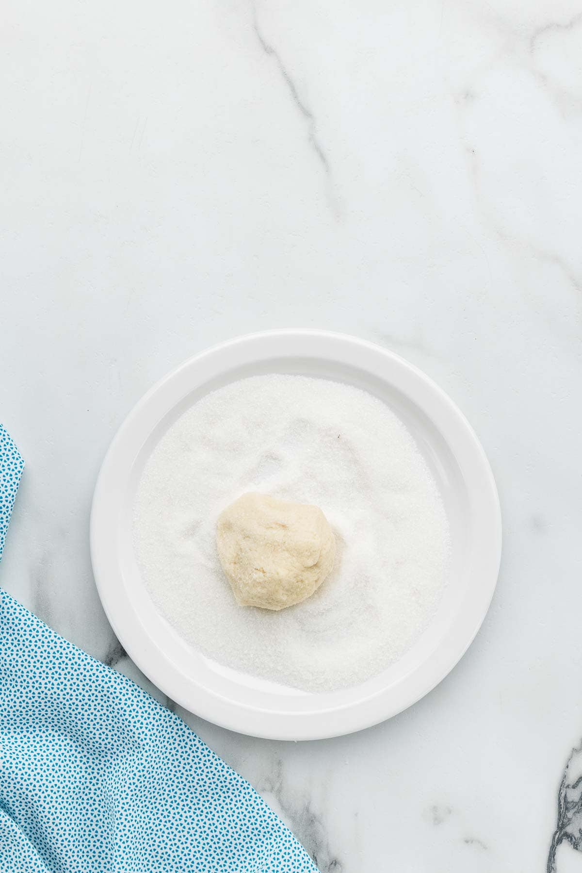 A ball of dough getting rolled in a small plate of sugar.