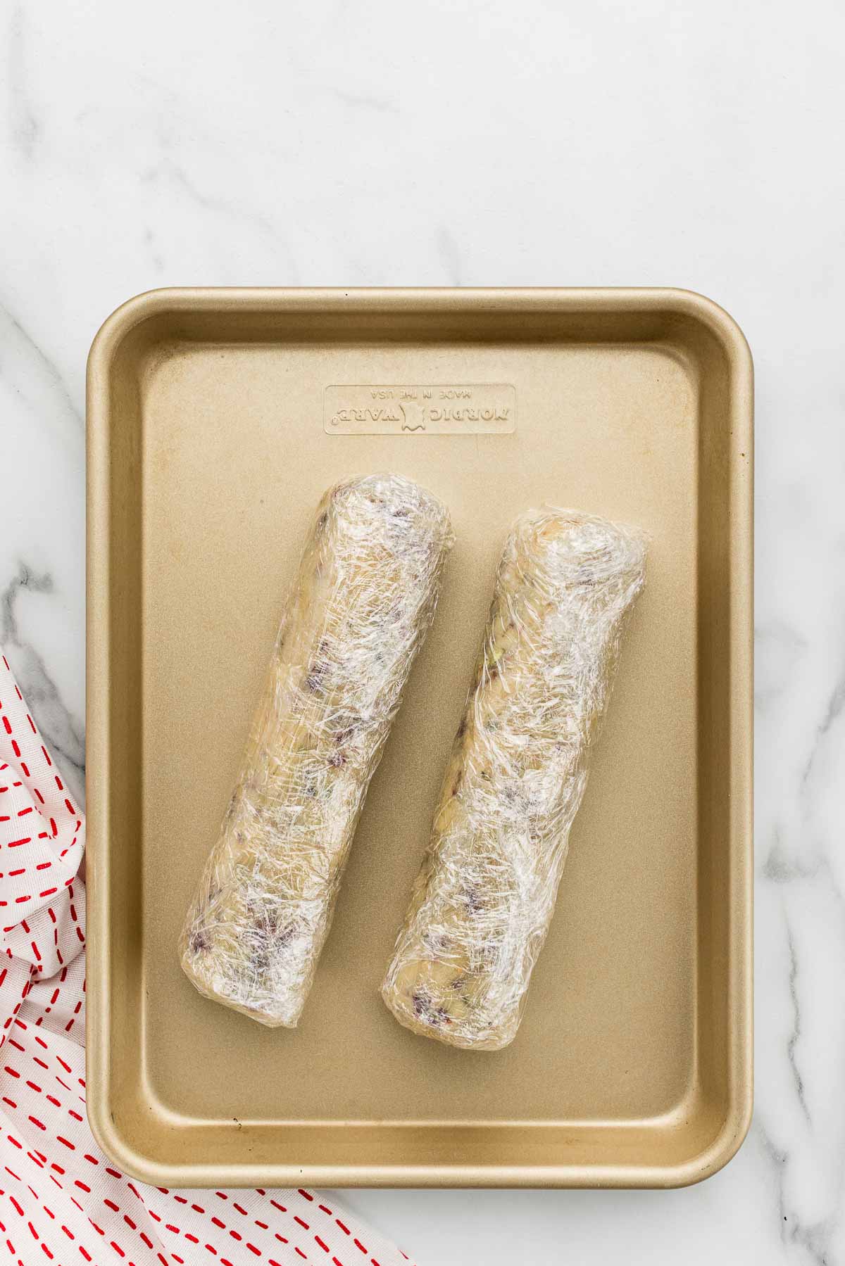 Pistachio cranberry cookie dough rolled into logs and wrapped on a sheet pan.