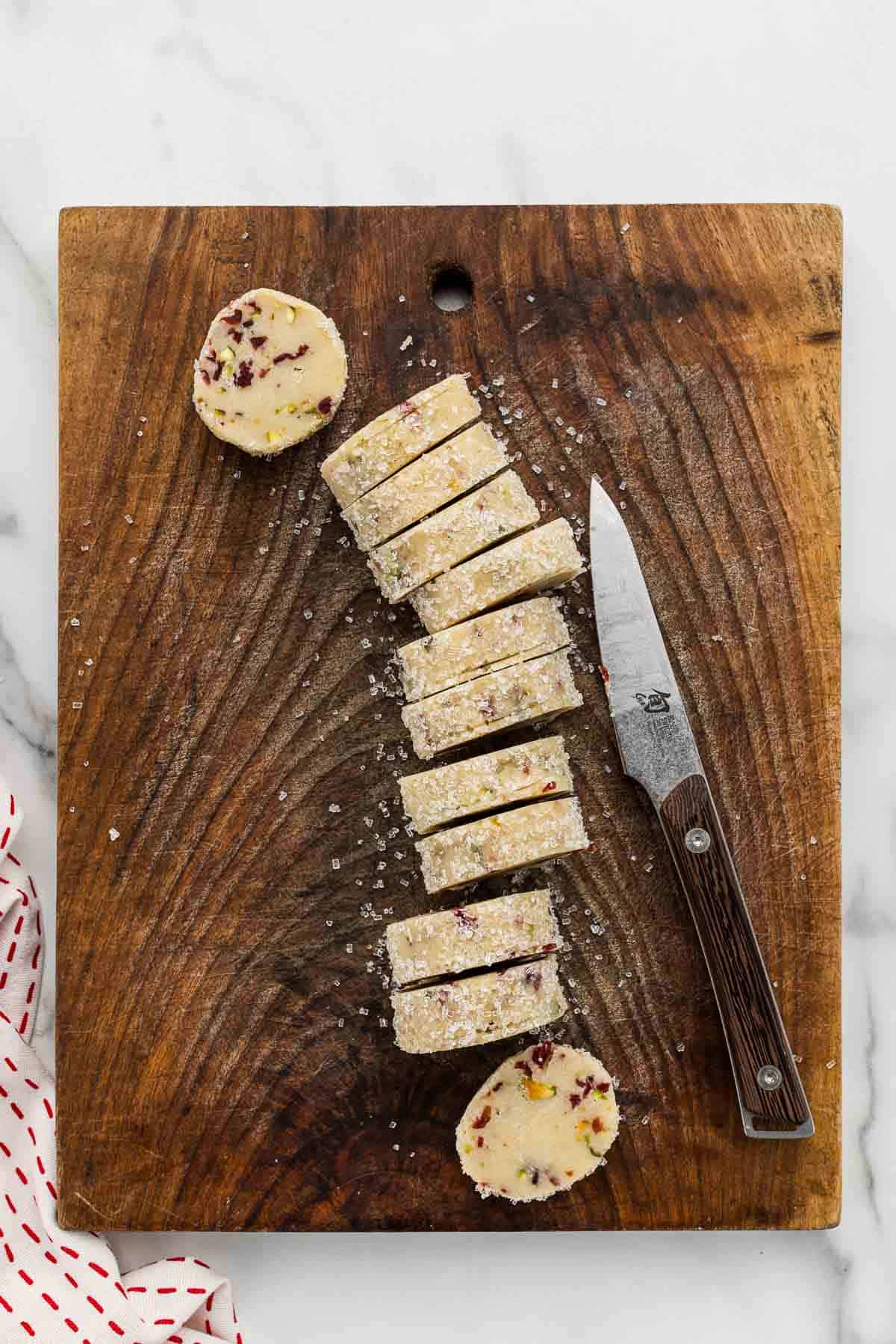 A wood cutting board with a sliced cookie dough log on it with a knife.