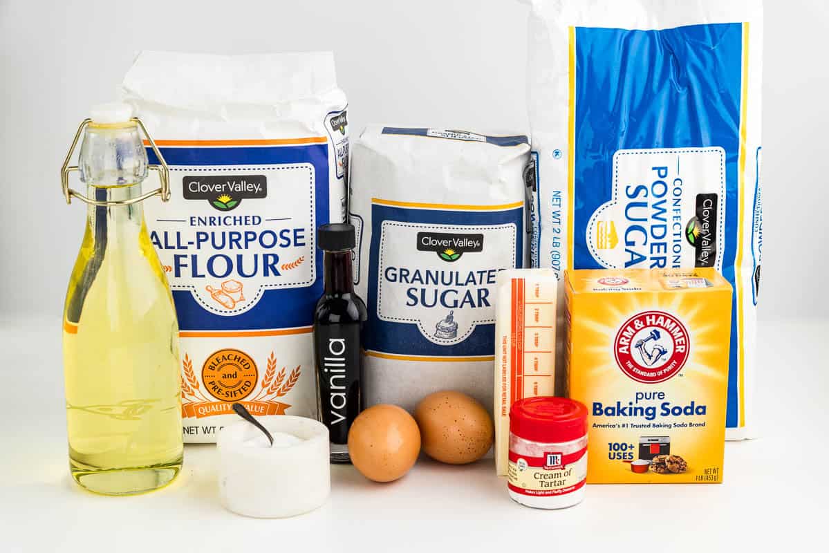 Oil, salt, flour, vanilla, eggs, sugar, cream of tarter, butter, baking soda, and powdered sugar on a counter.