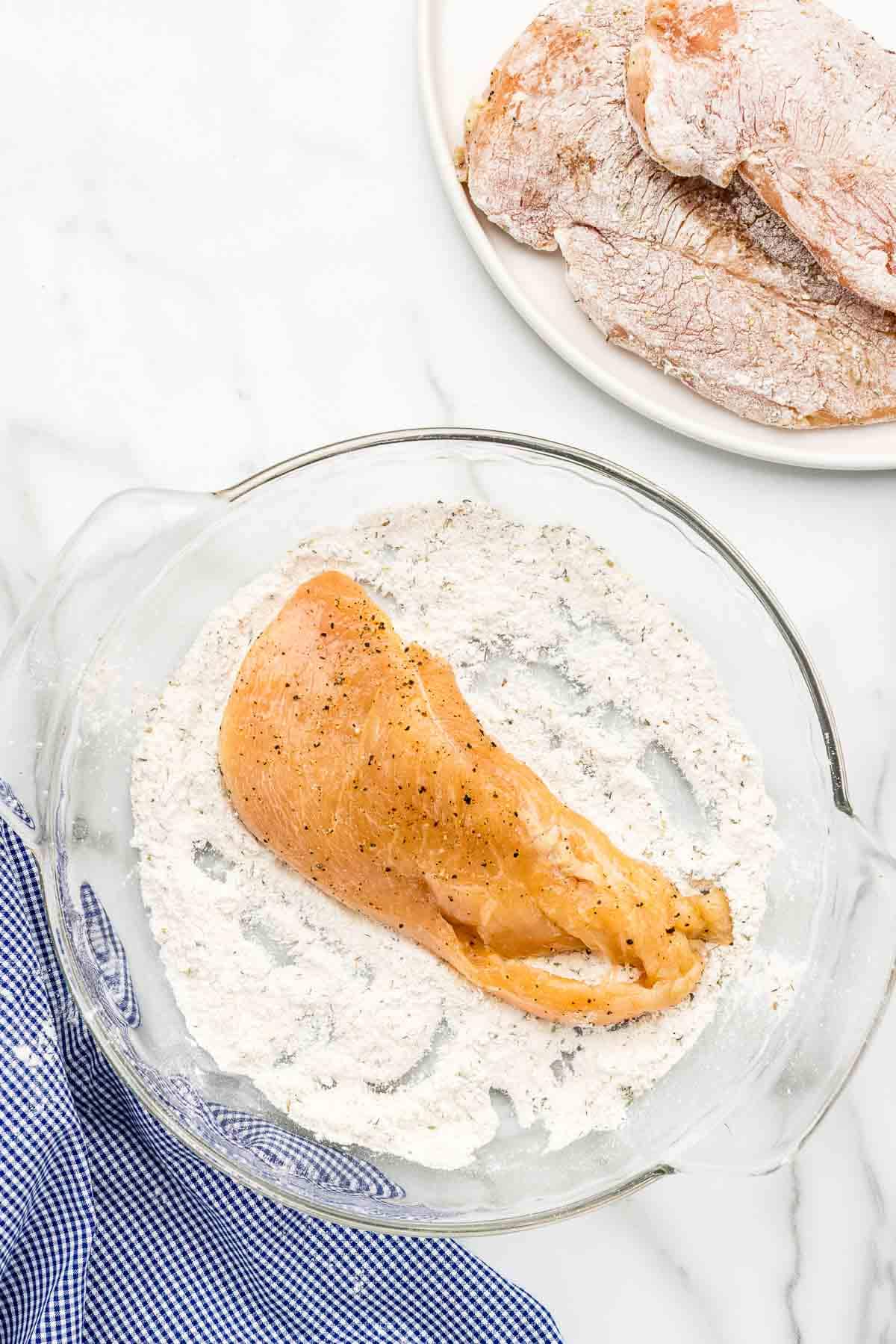 A raw chicken breast sitting in a glass dish of flour.
