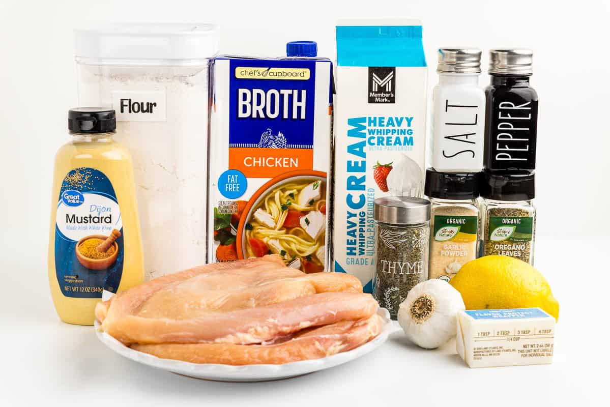 Dijon mustard, flour, chicken breasts, chicken broth, heavy cream, thyme, garlic, butter, garlic powder, oregano, salt and pepper, and a lemon on a counter.