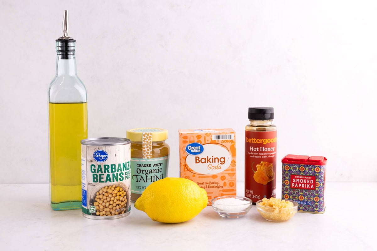 Garbanzo beans, oil, tahini, a lemon, baking soda, garlic, hot honey, and smoked paprika on a white surface.