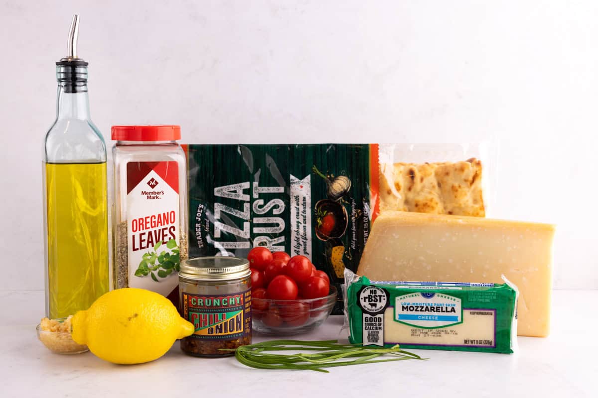 Herbs, cheese, flatbread, oil chili crisp, a lemon, tomatoes, and garlic on a countertop.