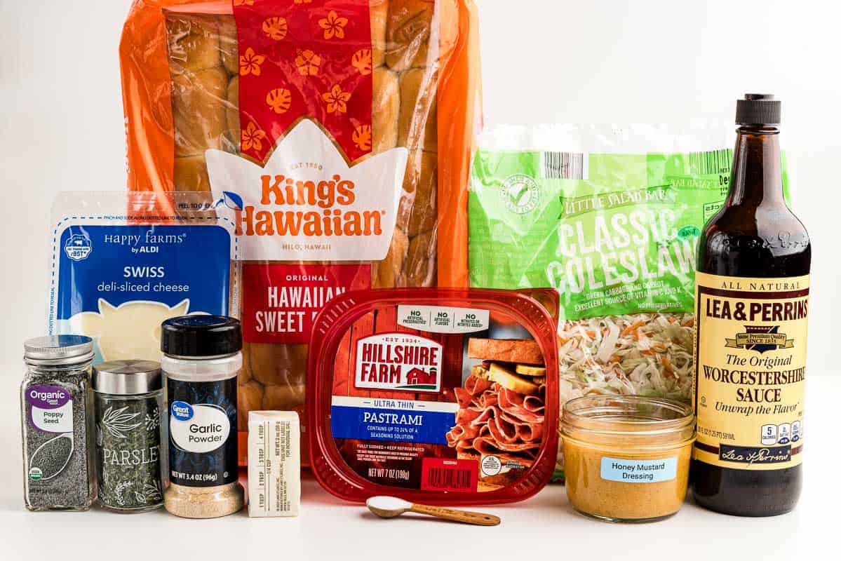 Cheese, Hawaiian rolls, pastrami, poppy seeds, parsley, garlic powder, butter, salt, honey mustard, Worcestershire sauce, and coleslaw on a white surface.