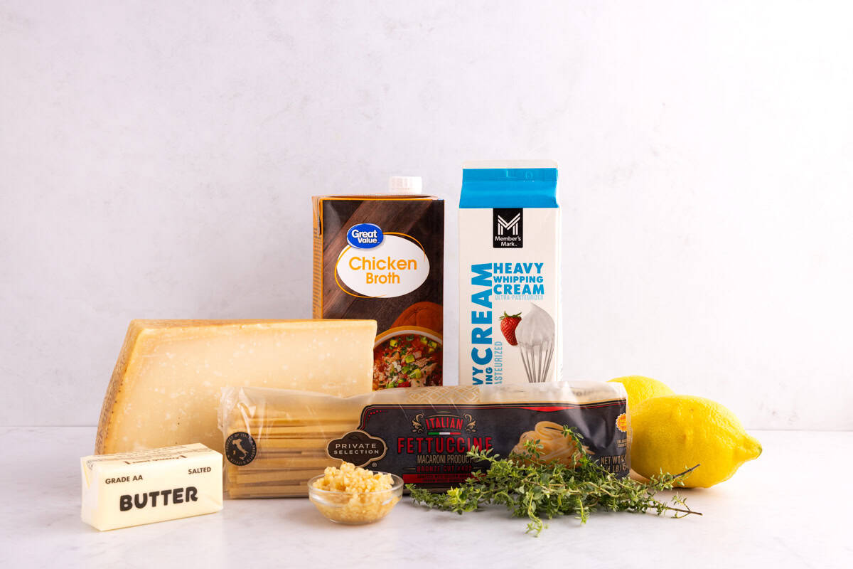 Butter, cheese, noodles, garlic, herbs, lemons, heavy cream, and chicken broth on a counter.