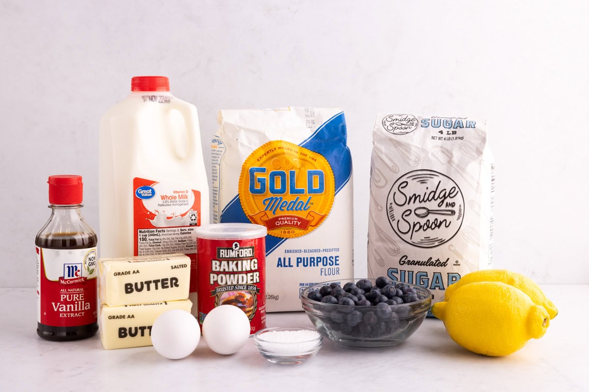 Vanilla extract, butter, eggs, milk, salt, baking powder, blueberries, flour, sugar, and lemons on a white surface.