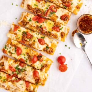 Flatbread pizza sliced on parchment with a small dish of toppings and a spoon beside it.