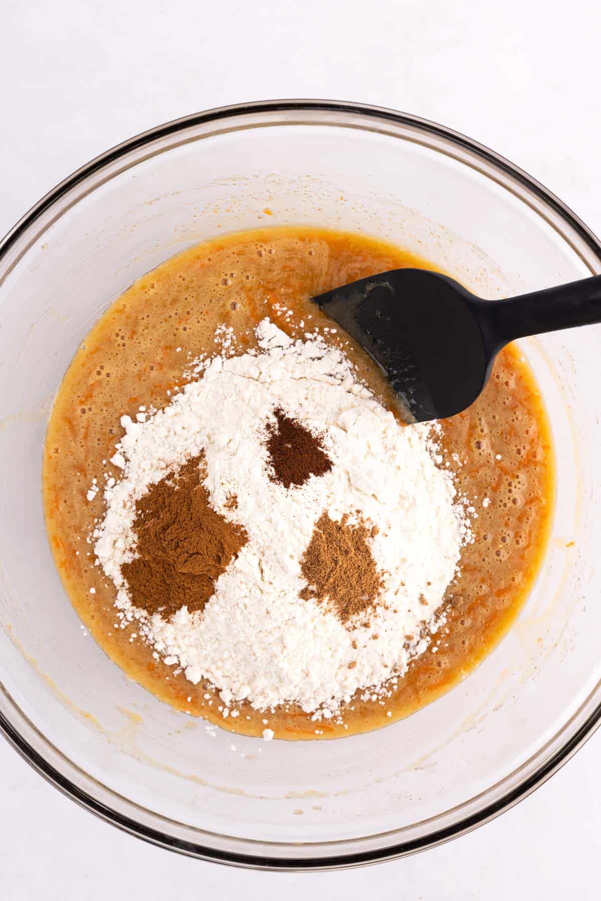 Dry ingredients added to carrot cake batter in a glass bowl with a rubber spatula.