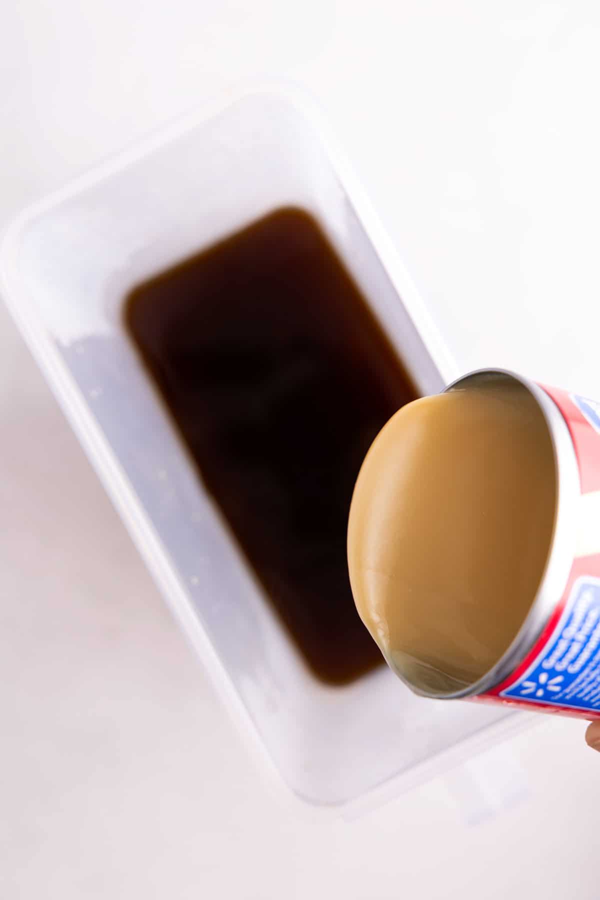 A can of condensed milk being poured in a plastic container of coffee.
