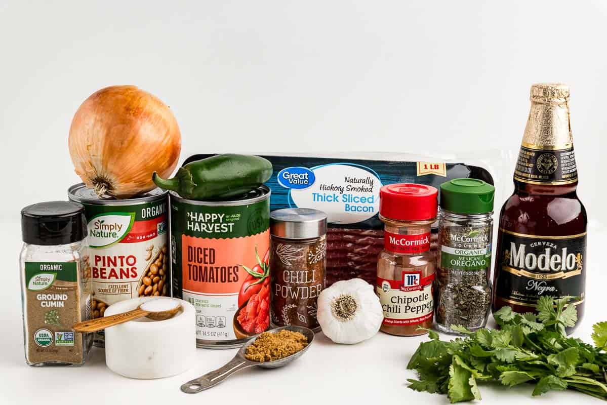 Seasonings, an onion, garlic, bacon, a jalapeno, cilantro, pinto beans, diced tomatoes, brown sugar, and beer on a countertop.