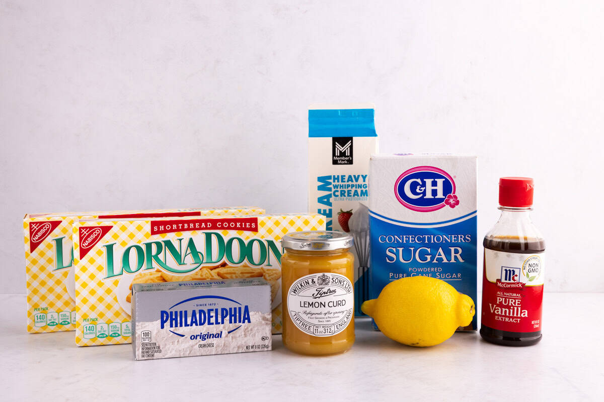 Shortbread cookies, cream cheese, lemon curd, a lemon, sugar, heavy cream, and vanilla extract on a white countertop.