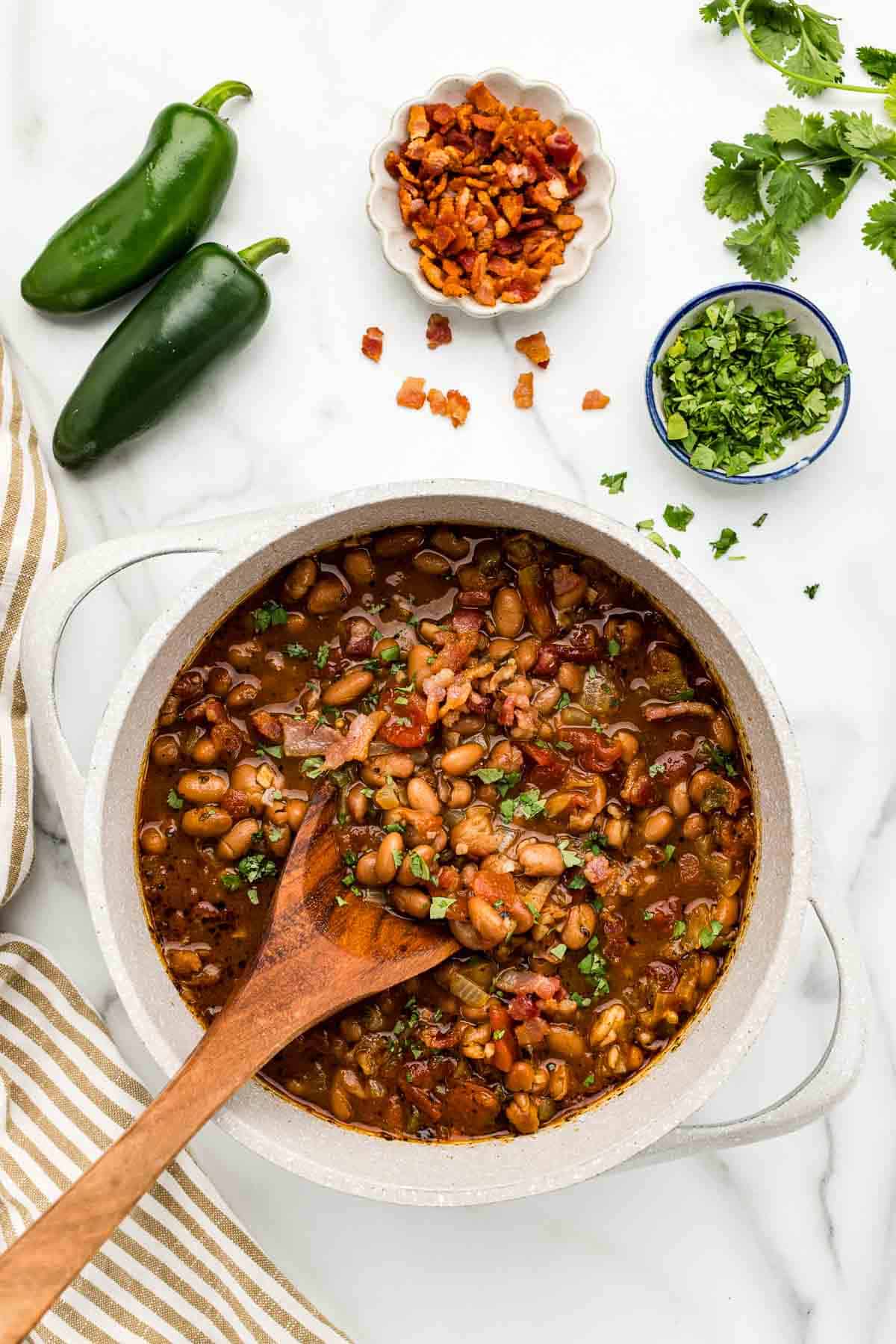 A large pot of borracho beans with a wooden spoon and an array of ingredients around it.