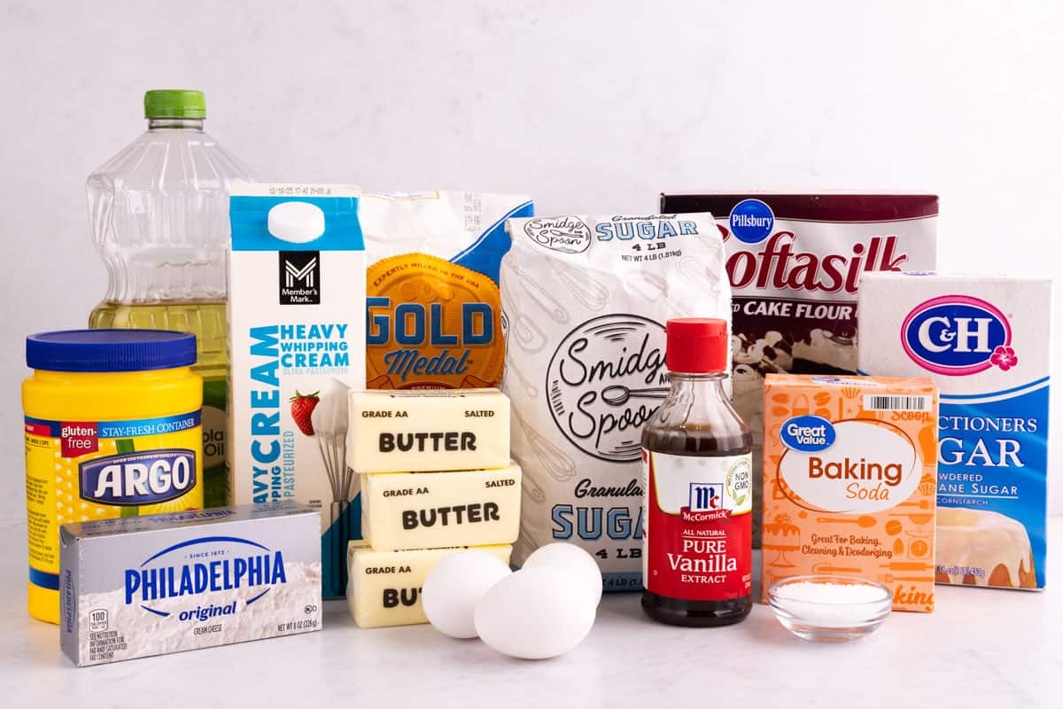 Corn starch, cream cheese, butter, oil, heavy cream, flour, eggs, sugar, vanilla extract, salt, baking soda, powdered sugar, and cake flour on a white surface.