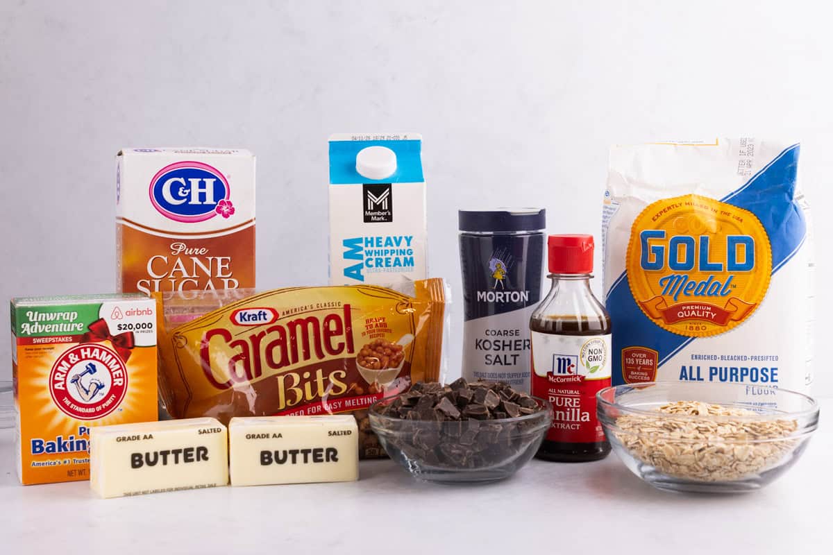 Baking soda, butter, caramel bits, cane sugar, heavy whipping cream, salt, vanilla extract, chocolate, flour, and oats on a countertop.
