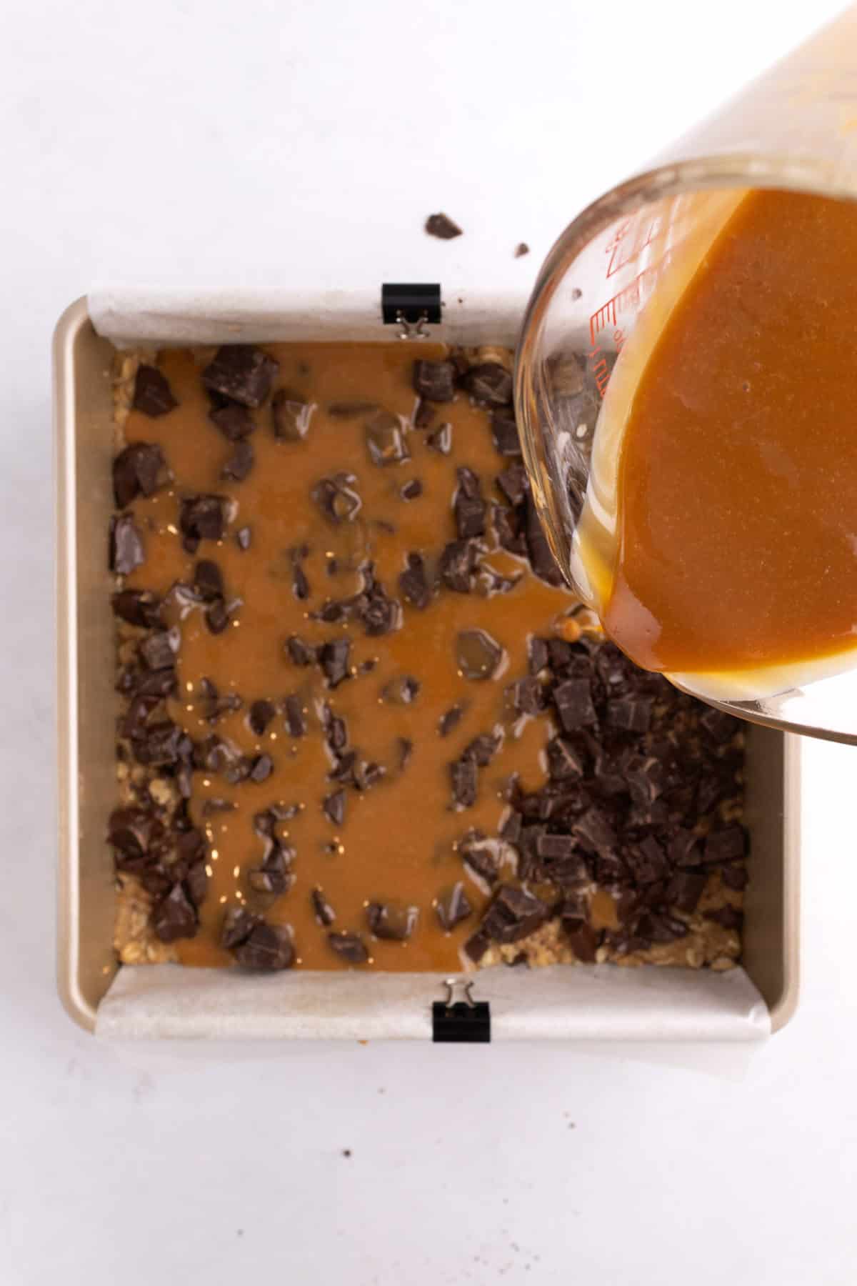 Caramel being poured into a pan with crust and chocolate chunks in it.