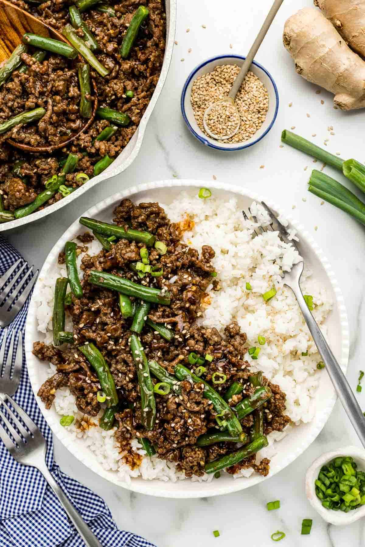 A serving of sesame ground beef over rice with an array of ingredients surrounding it.