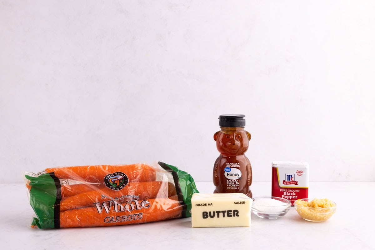 Carrots, butter, honey, salt, garlic, and black pepper on a white counter.