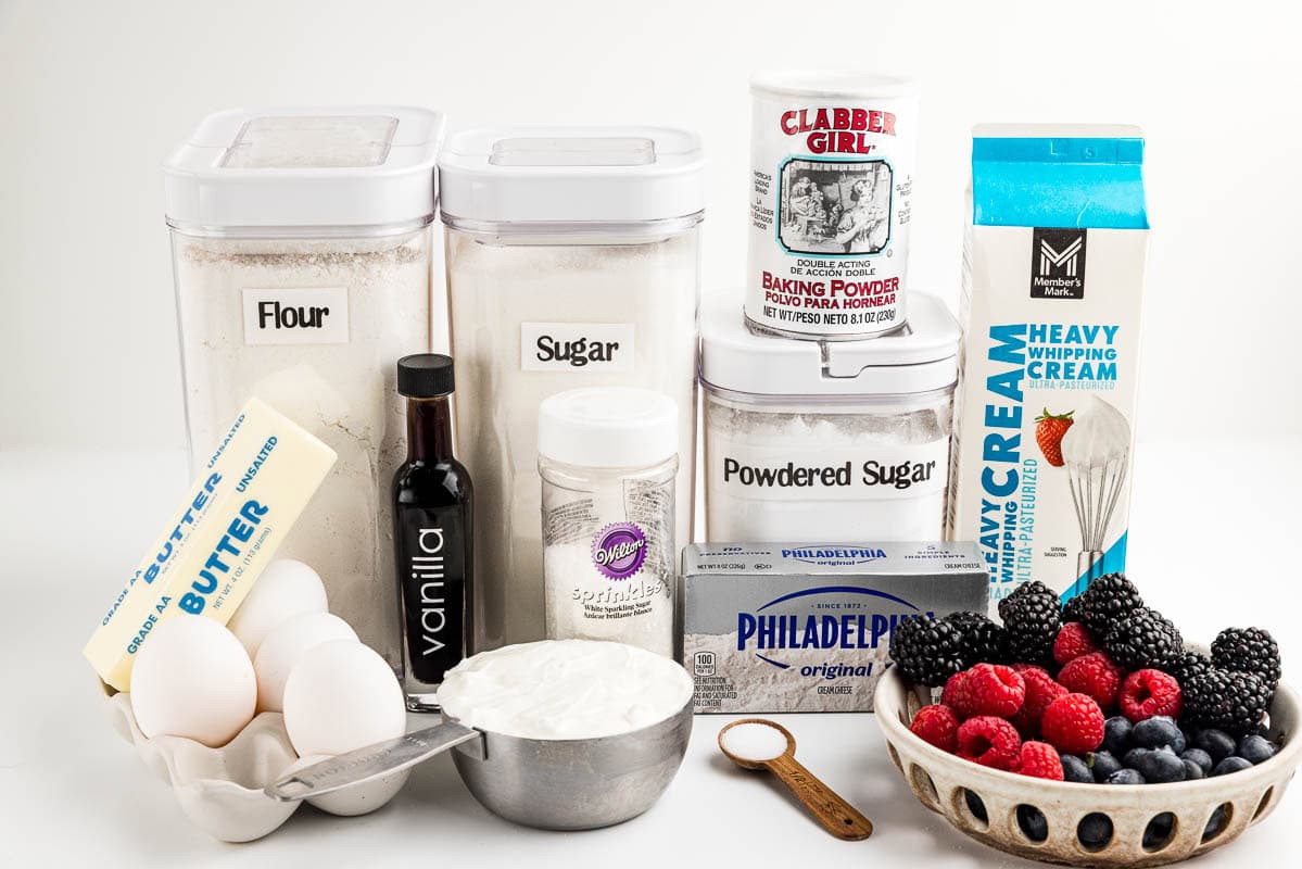 Flour, sugar, eggs, butter, powdered sugar, vanilla, sprinkles, salt, baking powder, heavy cream, fresh berries, cream cheese, and a cup of sour cream on a countertop.