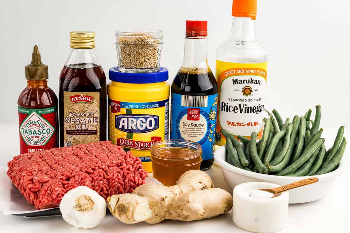 Hot sauce, sesame oil, corn starch, ground beef, garlic, ginger, sou sauce, salt, green beans, rice vinegar, sesame seeds, and honey on a counter.