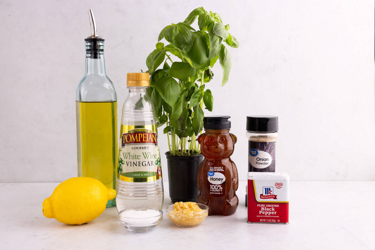 A lemon, garlic, salt, black pepper, onion powder, honey, basil, vinegar, and oil on a white surface.