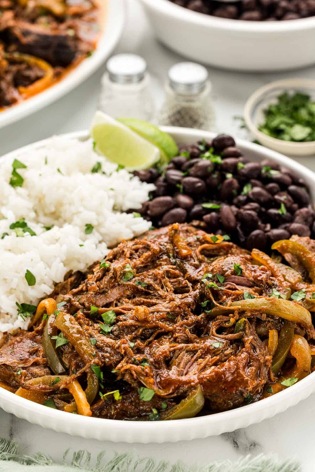 A plate of ropa vieja, beans, and rice with lime slices.